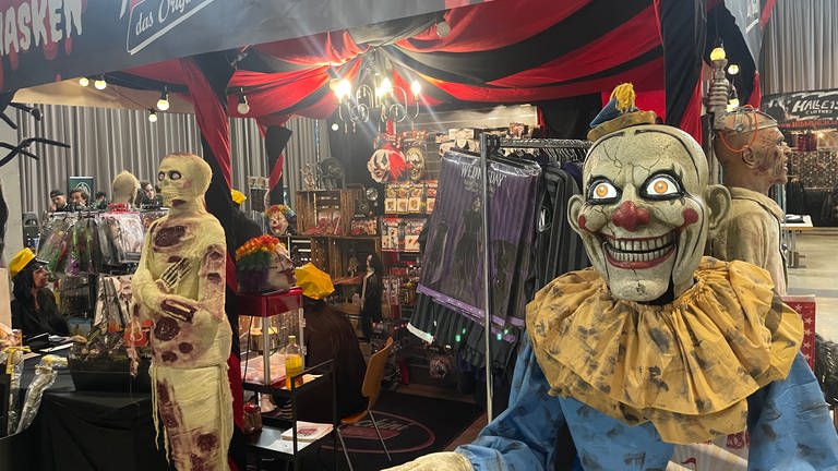 Ein Messestand mit Kostümen, davor eine gruselige Clown-Puppe und eine Mumie.